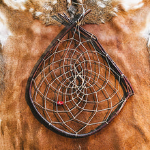 A handcrafted dreamcatcher with a dark brown wooden frame, white webbing, and a small red bead, displayed on a brown animal hide background.