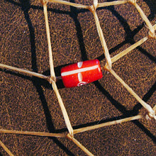 A red cylindrical bead with a white cross design, threaded on a natural tan string against a dark brown textured background.