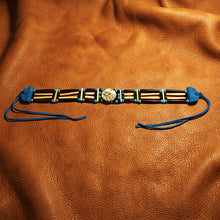 A beaded bracelet laid flat on a textured brown leather background. The bracelet features dark brown and light tan wooden beads, turquoise blue cord accents, and a central round silver-toned medallion, with blue cord ties extending on both ends.