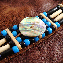 A beaded bracelet featuring a large iridescent mother-of-pearl shell centerpiece, bright blue beads, black beads, and light natural wood beads, displayed on a brown textured leather background.