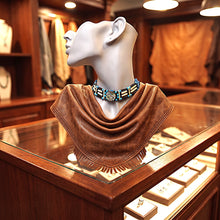 A brown draped scarf or shawl with fringe, displayed on a white mannequin head in a jewelry store. The mannequin is wearing a turquoise and black beaded collar necklace.