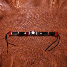 A beaded bracelet laid flat on a textured brown leather background. The bracelet features multiple strands of black beads, red and white woven accents, small metallic silver beads, and a central white circular bead, with red end caps and black hanging ties.