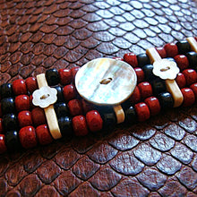 A beaded bracelet featuring red and black beads, light wood accents, a white flower-shaped bead, and a large round mother-of-pearl button, displayed on a brown textured leather background.