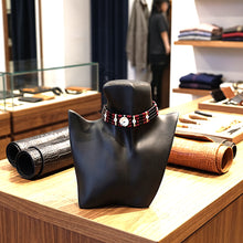 A beaded choker necklace with a central white oval stone, displayed on a black mannequin bust inside a jewelry or leather store. Rolled leather pieces and wooden shelves are visible in the background.